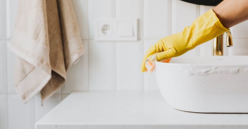 Cleanser - Crop anonymous person wearing yellow latex gloves washing sink in bathroom with orange sponge and cleanser while cleaning contemporary flat