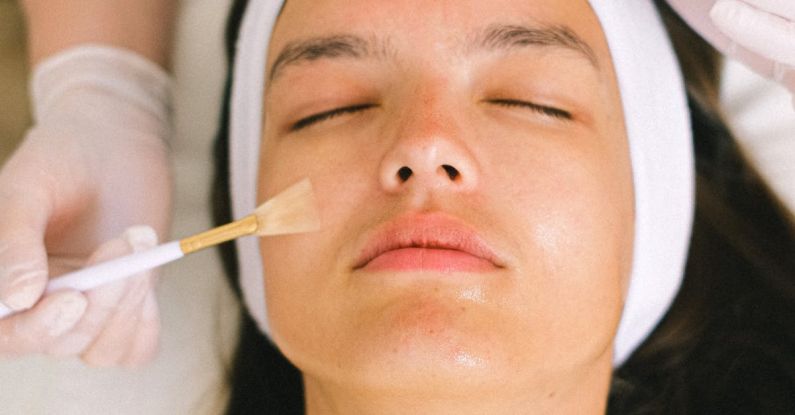 Serums - Top view skilled female cosmetologist in uniform and gloves applying essential oil on young woman face skin while working in contemporary spa center