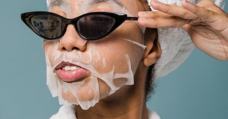 Moisturizers - Self esteem young African American lady with moisturizing sheet mask and towel on hand wearing trendy sunglasses while standing against blue background in bathrobe after shower